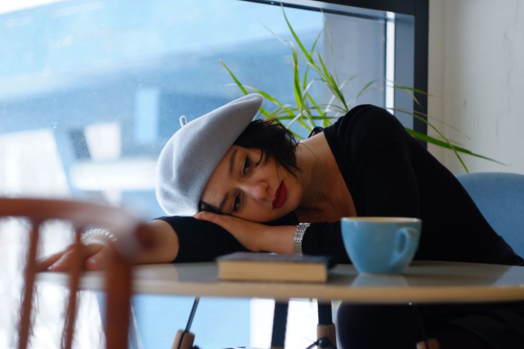 Woman in blue tam leaning on her arm with blue teacup and book, looking sad. Photo by Aleksandra Sapozhnikova on unsplash.com